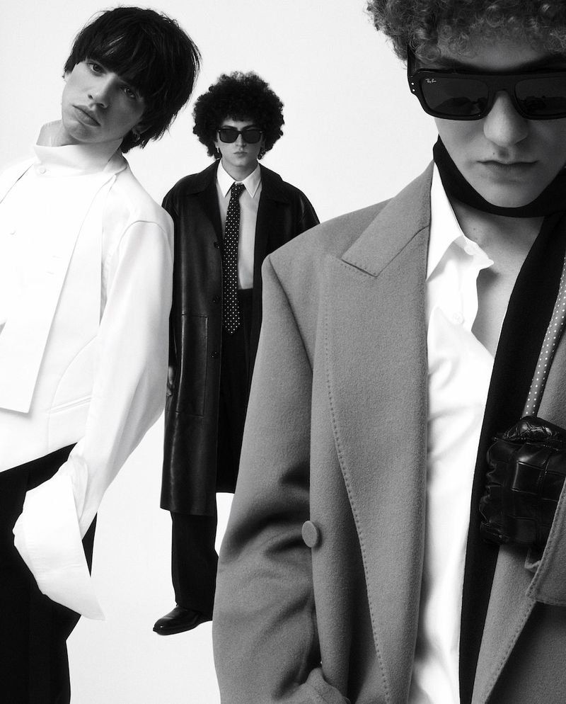 Three men pose in a monochrome fashion shot. One wears a white suit, another a leather coat, and the third a grey blazer with sunglasses.