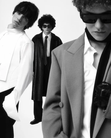 Three men pose in a monochrome fashion shot. One wears a white suit, another a leather coat, and the third a grey blazer with sunglasses.