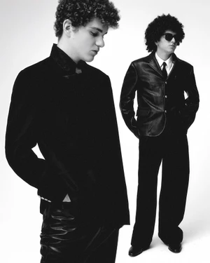 Two male models posing in tailored black outerwear against a bright white studio backdrop.