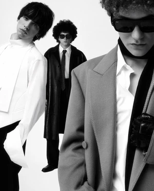 Three young men posing in tailored menswear including a grey blazer, black leather trench coat, and white structured shirt on white backdrop.
