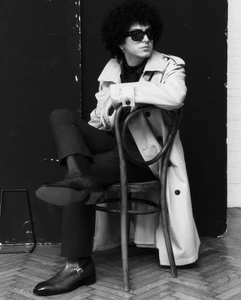 Black and white portrait of a person with curly hair wearing dark sunglasses, a trench coat, and dress boots on a wooden chair.