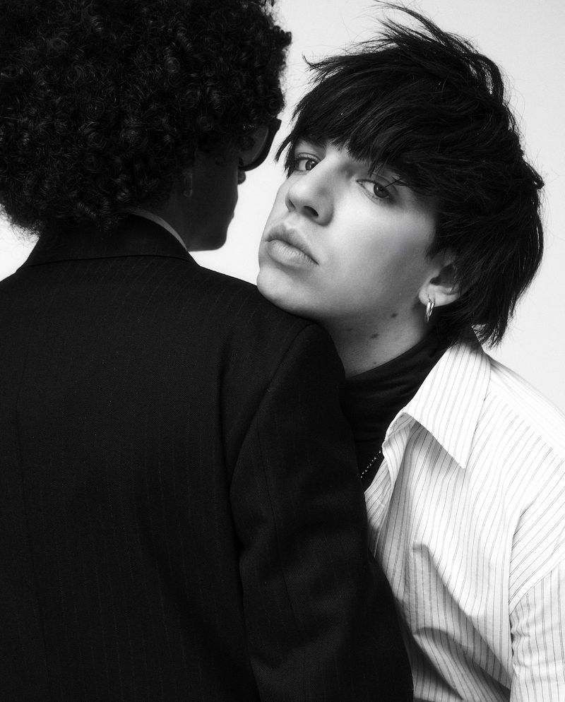 Two people posing intimately, one wearing a pinstripe shirt resting head on another in a black suit