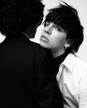 Two people posing intimately, one wearing a pinstripe shirt resting head on another in a black suit