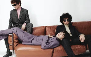 Three young men in dark suits and sunglasses posing with a brown leather couch in a fashion editorial shoot.