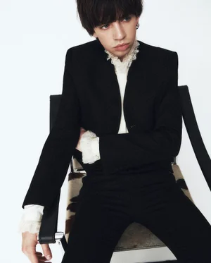Model wearing a tailored black suit with a white ruffled lace collar and cuffs seated in a chair against a white background.