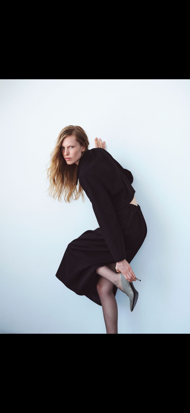 Model Leanne De Haan wearing a black long-sleeve dress and pointed mules against a plain white wall.