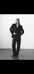 Model Leanne De Haan in a black tailored suit with an oversized blazer, cropped vest, and wide-leg trousers in a white studio setting.