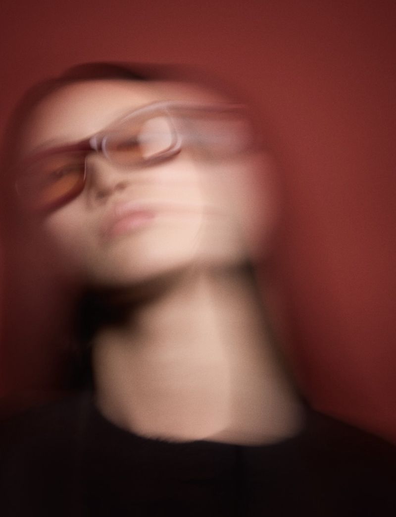Blurred portrait of a woman with glasses against a red background, creating a sense of motion and abstraction in a close-up shot.