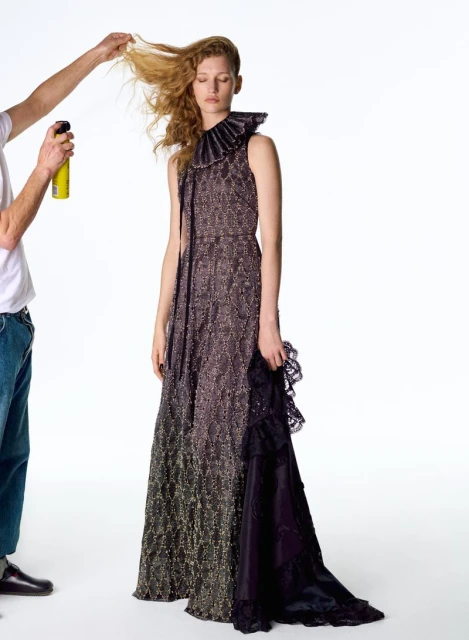 A fashion model poses in a formal, shimmering dress with a ruffled collar, while a stylist works on her hair against a white background.