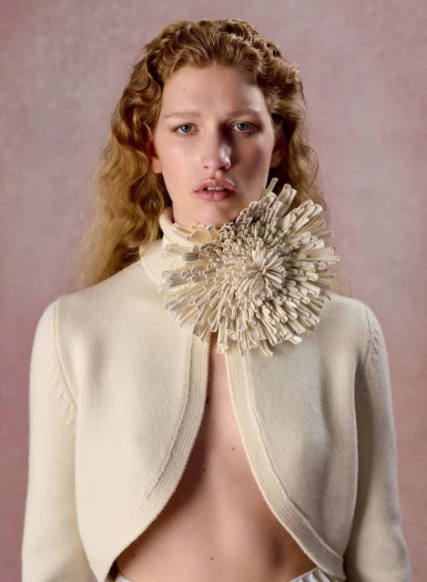 Blonde model poses in front of a pink background, wearing an off-white sweater and a floral collar in a studio fashion shot.