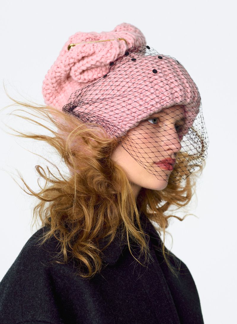Woman in a pink knitted beret with a black dotted net veil, posing in a photo studio with a simple white background.