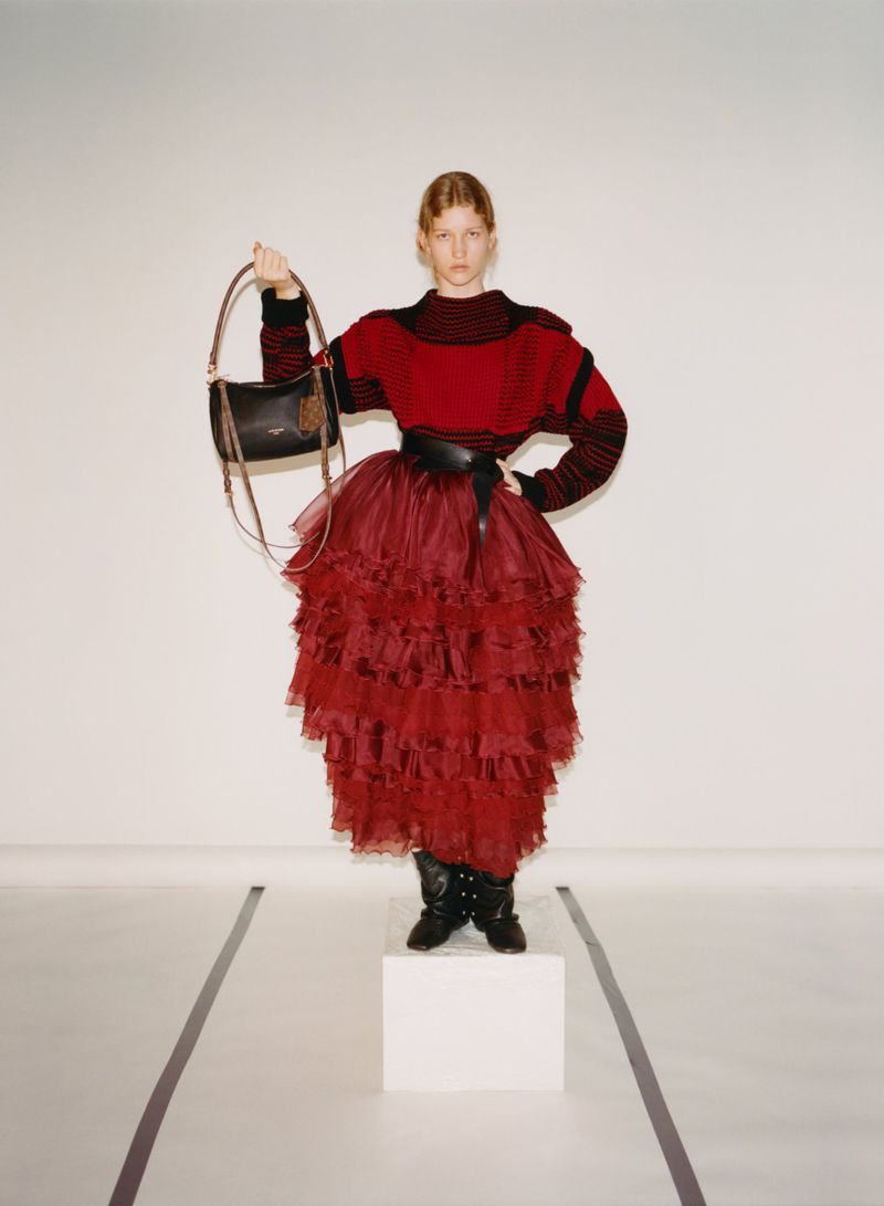 Adult female fashion model in a red sweater and layered tulle skirt poses in a photo studio holding a black shoulder bag.