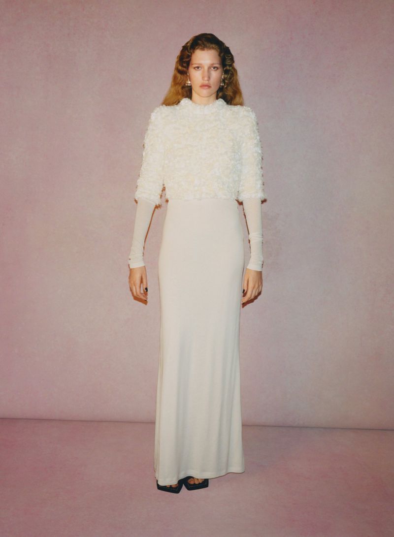 Full-length studio shot of a young woman in a cream-colored dress and cropped top, posing in front of a faded pink backdrop.