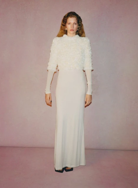 Full-length studio shot of a young woman in a cream-colored dress and cropped top, posing in front of a faded pink backdrop.