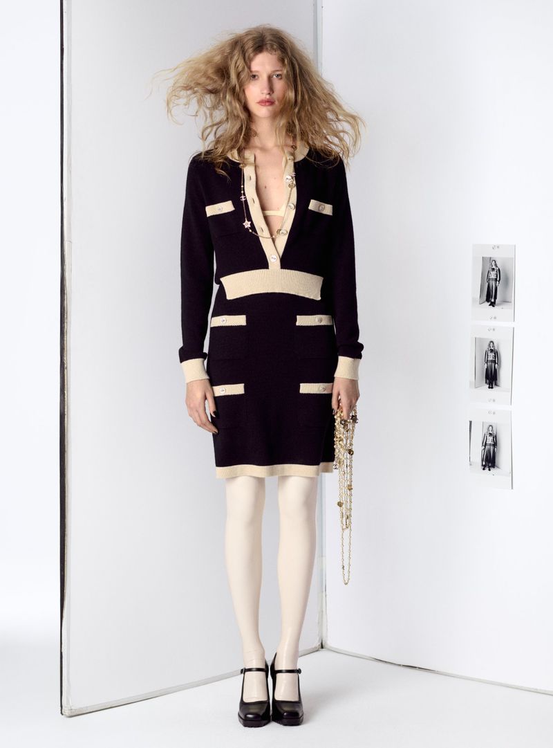 Woman poses in a black and beige collared dress with stockings and Mary Jane shoes in a white corner studio fashion shot.