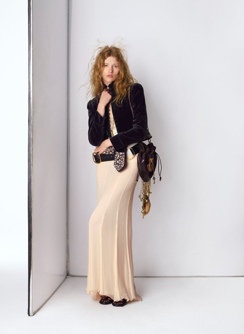 Fashion shot of a woman in a dark velvet jacket and long dress posing in a photo studio with a light reflector visible in the background.