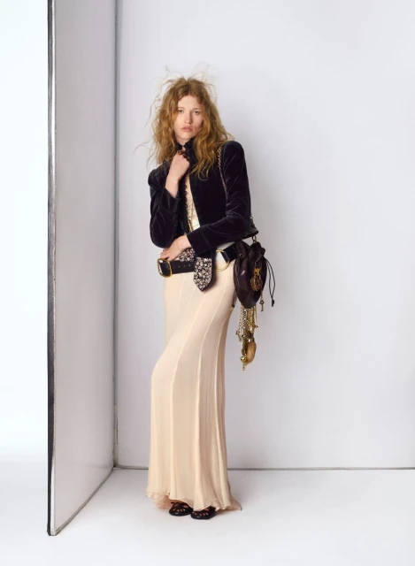 Fashion shot of a woman in a dark velvet jacket and long dress posing in a photo studio with a light reflector visible in the background.
