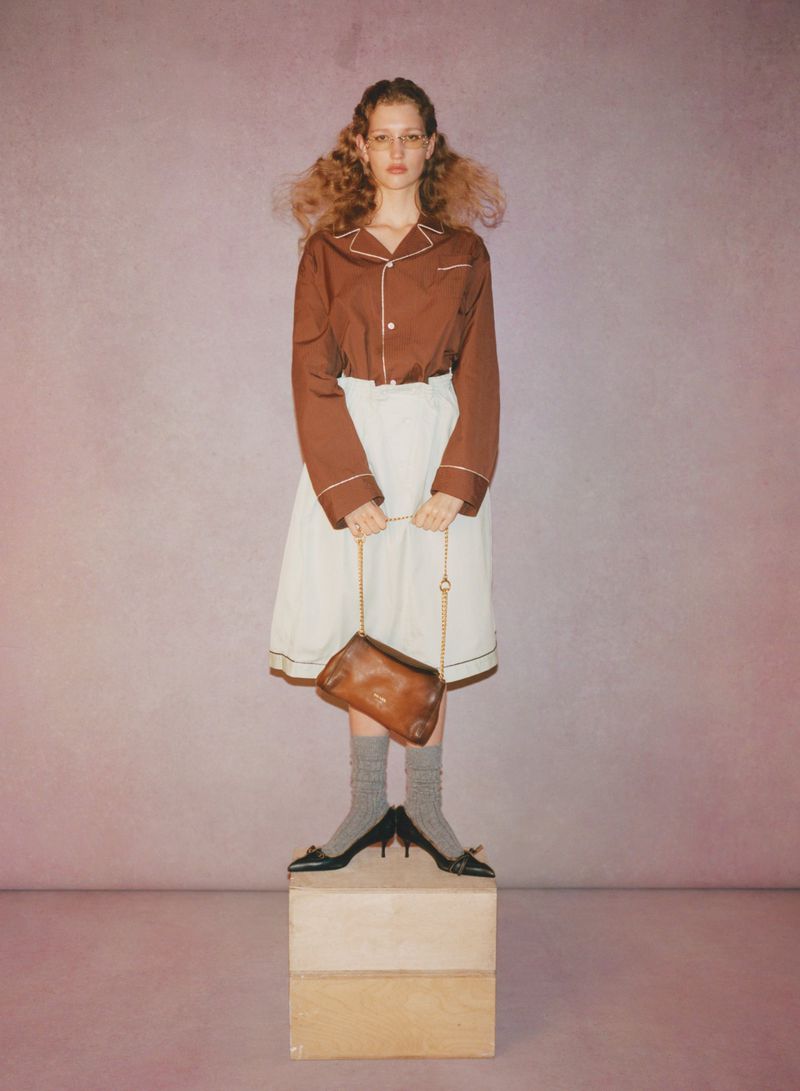 Full-length portrait of an adult female with voluminous hair and a light complexion, standing on boxes against a soft pink background.