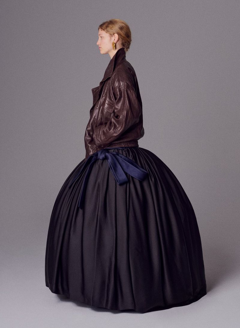 Profile view of an adult female fashion model in a brown leather jacket and large black formal gown with blue ribbon in a studio.