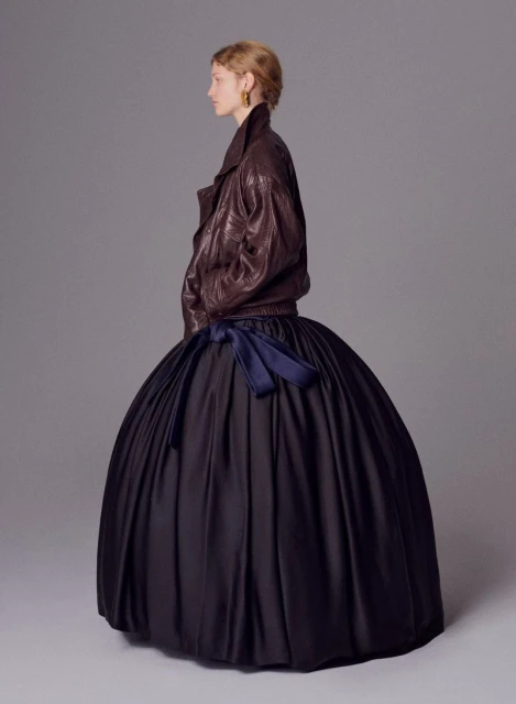 Profile view of an adult female fashion model in a brown leather jacket and large black formal gown with blue ribbon in a studio.