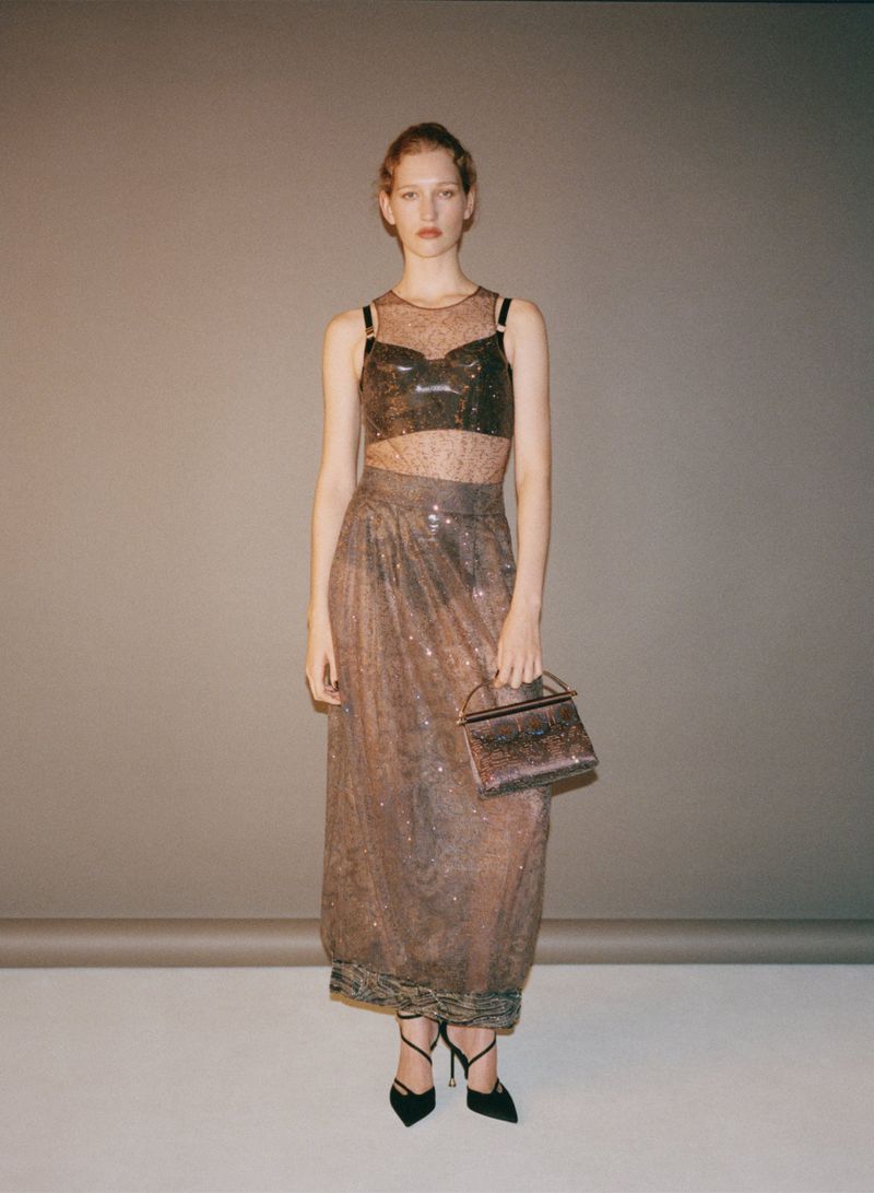 Full-length studio shot of a young woman in a shimmering, semi-sheer two-piece outfit and heels, holding a boxy handbag.