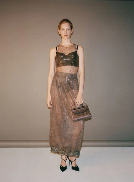 Full-length studio shot of a young woman in a shimmering, semi-sheer two-piece outfit and heels, holding a boxy handbag.