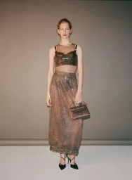 Model Apolline Rocco Fohrer posing in a sheer embellished dress and black heels while holding a handbag against a studio backdrop.
