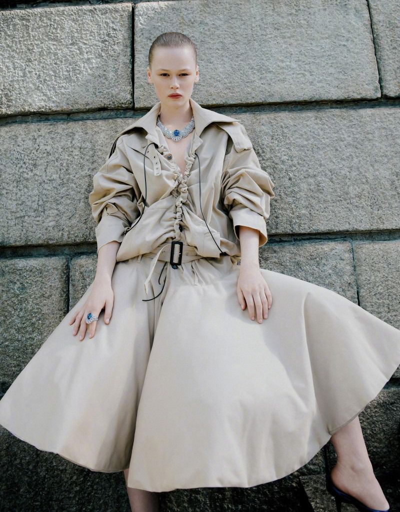 Fashion shot of a woman in a beige outfit with jewelry posing against a stone wall in an outdoor setting, captured in natural light.