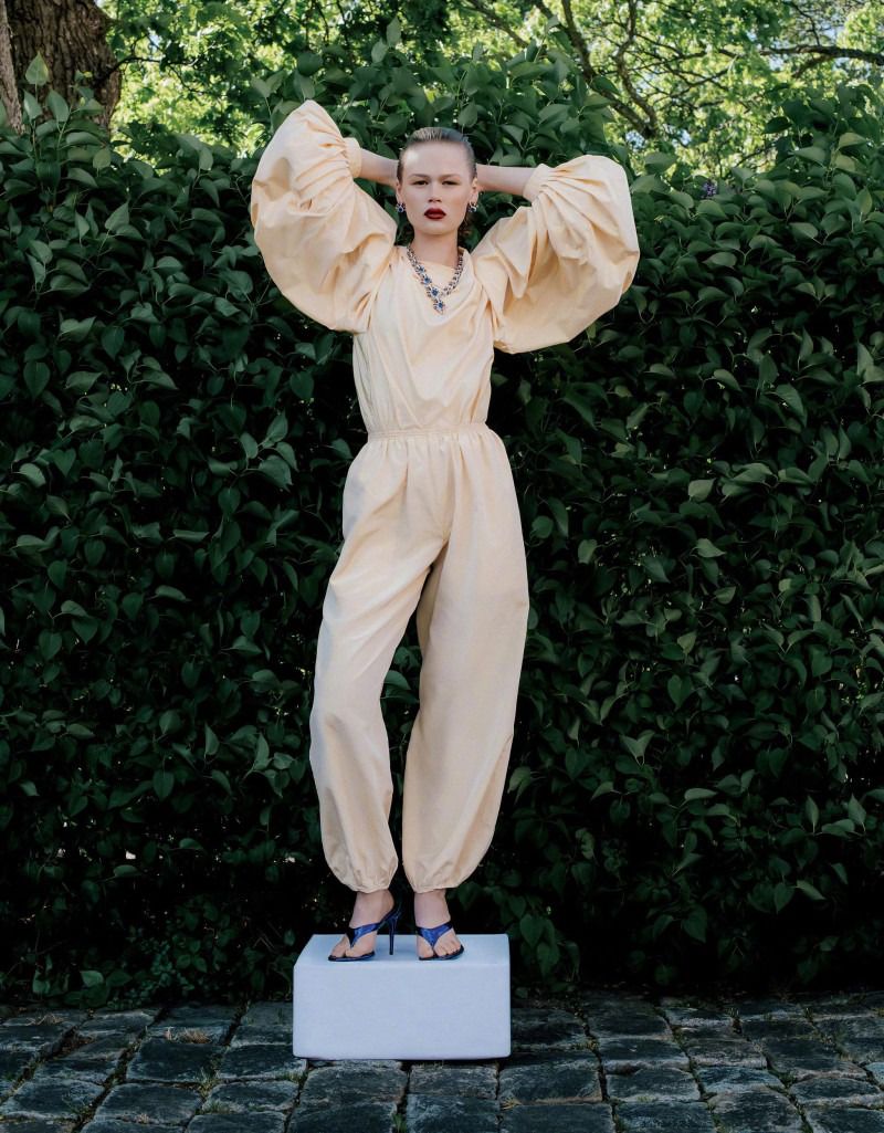 A woman with red lipstick, wearing an oversize cream-colored jumpsuit and heels, poses against a green hedge in an outdoor setting.