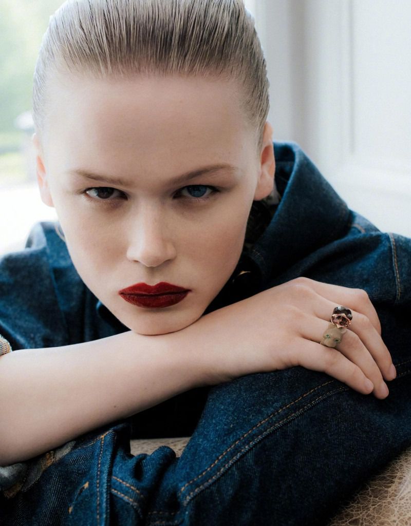 A portrait of a young woman with fair skin and dark red lipstick, wearing a blue denim jacket and a large ring, resting her chin on her hands.