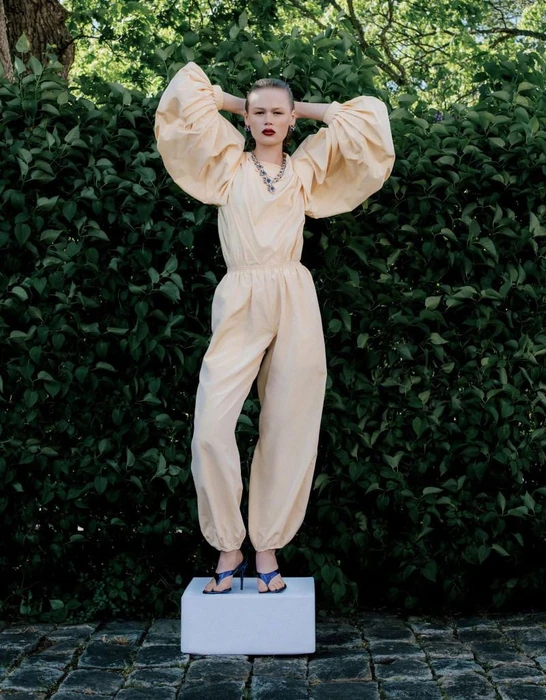 Model wearing a pale yellow voluminous jumpsuit standing on a white block against dense green foliage.