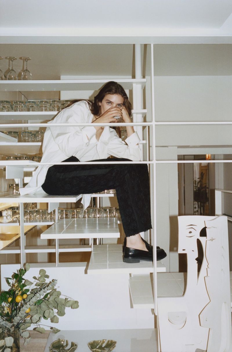 A young woman sits on a white staircase in a casual white shirt and black trousers, with decorative glassware and modern decor.