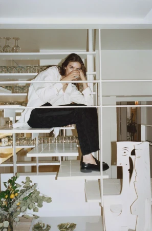 A young woman sits on a white staircase in a casual white shirt and black trousers, with decorative glassware and modern decor.