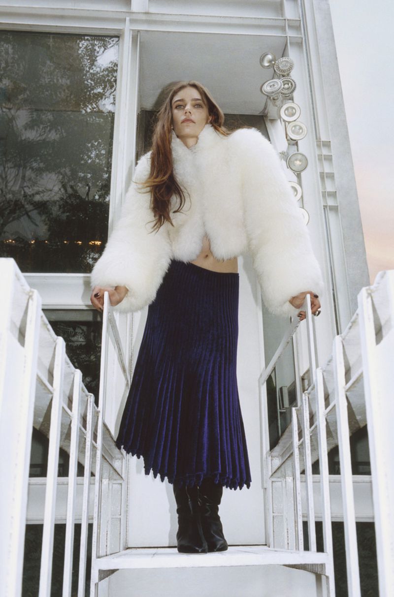A full shot of a woman in a white coat and blue skirt standing on white metal stairs outside during the day.