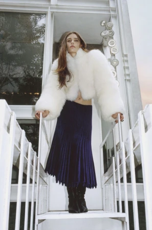 A full shot of a woman in a white coat and blue skirt standing on white metal stairs outside during the day.