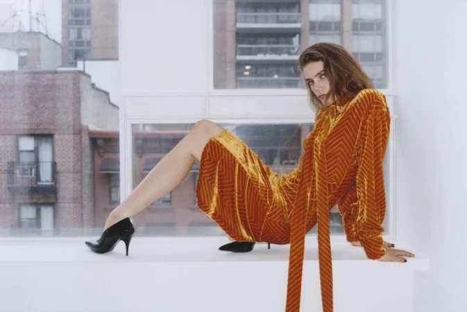 A woman with long brown hair poses indoors sitting on a windowsill, wearing a long-sleeved, textured amber dress and black high heels.