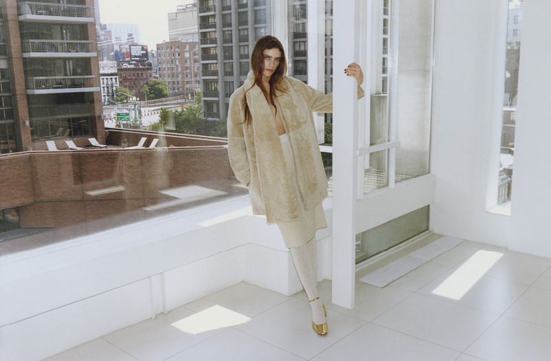 Fashion shot of a woman in a beige coat, beige skirt, and gold shoes, posing inside a high-rise apartment with a city view.