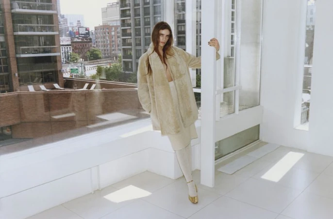 Fashion shot of a woman in a beige coat, beige skirt, and gold shoes, posing inside a high-rise apartment with a city view.
