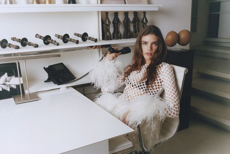 Adult woman seated in chair at desk with telephone, wearing white feathered dress in an indoor environment with unique decor.