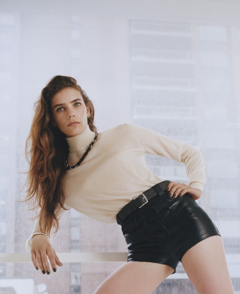 Fashion photo of a slender woman with long wavy hair, wearing a beige turtleneck, black shorts, and a chain necklace, posing indoors.