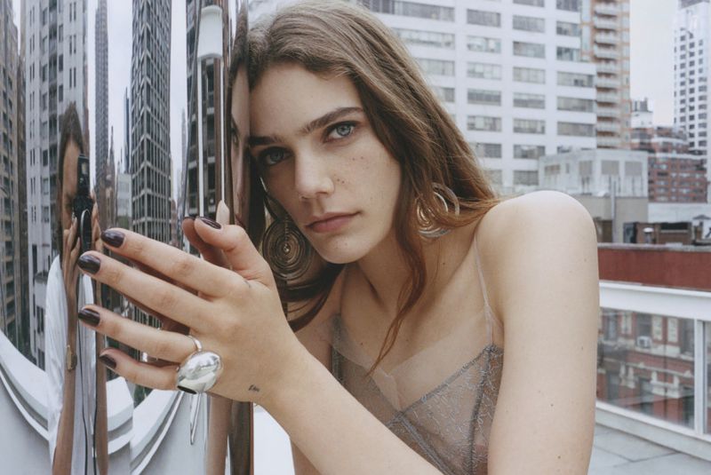 Portrait of a young woman with long brown hair, wearing sheer top and hoop earrings, posing with a reflective building in the background.