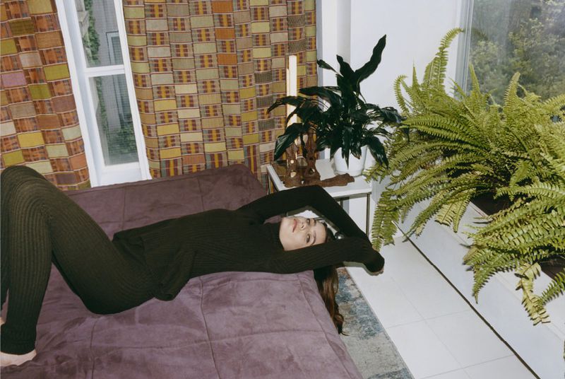 Adult woman lying on a purple couch in a casual outfit next to indoor houseplants, a patterned wall, and a large window.