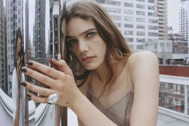 Model leaning against a mirrored chrome pillar with a camera reflection and a city skyline background.