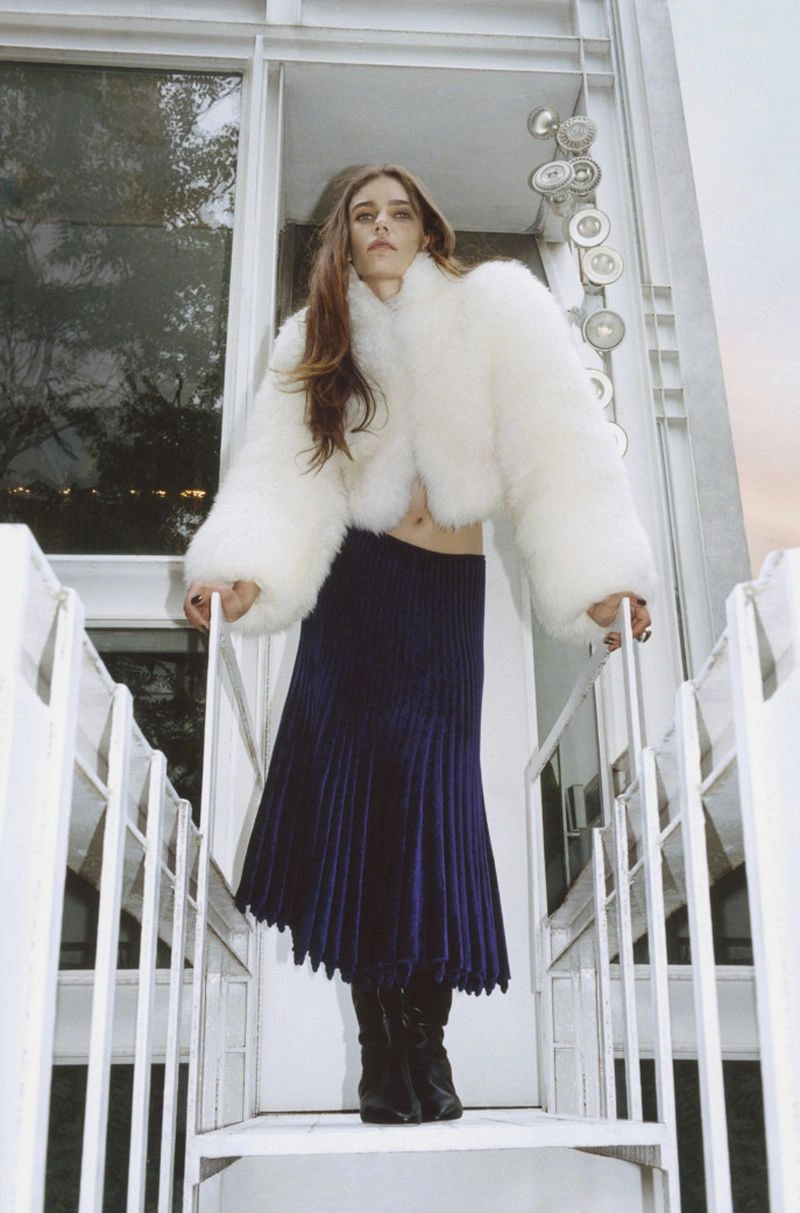 Model Betsy Gaghan poses on an exterior white staircase wearing a cropped white faux-fur jacket, navy pleated skirt, and black boots.