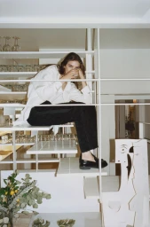 Model seated on white modern stairs wearing a white shirt and black trousers, holding a glass for Elle U.S. Modern Love project.