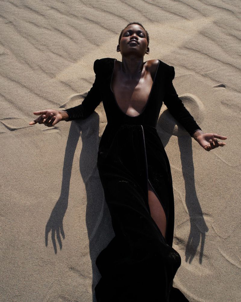 A black model in a dark dress with a plunging neckline lies in the sand with her arms outstretched, casting a long shadow on the dunes.