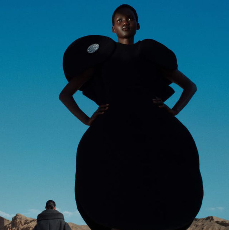 Adult female fashion model poses in a dark avant-garde gown outdoors against a clear blue sky with a man in the distance.