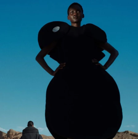 Adult female fashion model poses in a dark avant-garde gown outdoors against a clear blue sky with a man in the distance.