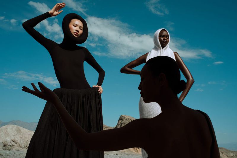 Three fashion models in stylized dresses and headwear pose in a desert landscape under a bright blue sky with some clouds.