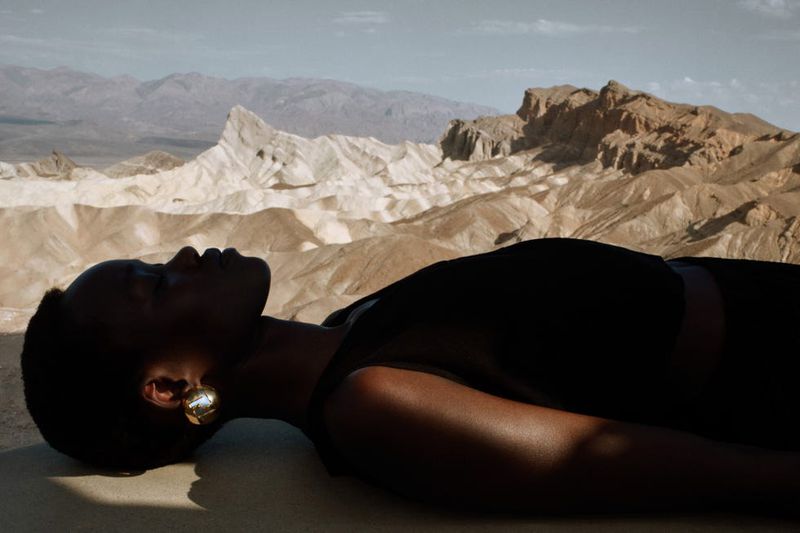 An African-descent woman in a sleeveless top lies on tan surface against a desert backdrop, illuminated by natural light, wearing gold earrings.